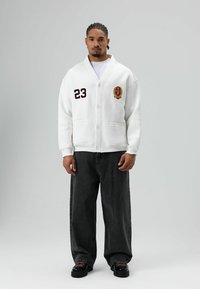 Witte cardigan met een sjaalkraag, voorzien van een geborduurd bordeauxkleurig nummer "23" en logo, gecombineerd met donkere, wijde jeans en zwarte schoenen.