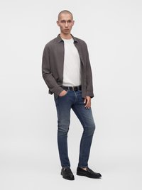 Homme portant un t-shirt blanc, une chemise gris foncé déboutonnée, un jean bleu, une ceinture noire et des mocassins noirs, debout avec une main dans la poche.