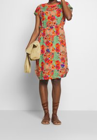 Das Blumenkleid hat kurze Ärmel, einen elastischen Bund und mehrfarbige Blumen auf einem pfirsichfarbenen Hintergrund. Kombiniert mit braunen Riemchensandalen.