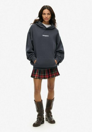 Dunkelgrauer oversized Hoodie mit einer Fronttasche und Logo, kombiniert mit einem rot-, grün- und gelb-karierten Faltenrock und braunen knielangen Stiefeln.
