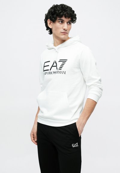 Бяла суитшърт с качулка и преден джоб, с черно лого "EA7 Emporio Armani"; комбинирана с черен панталон с малко лого.