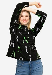 Schwarzes Langarmshirt mit großen, weißen und grünen Buchstabenprints. In Kombination mit blauen Jeans hat das Shirt einen lockeren Schnitt.