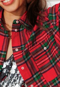 Chemise à carreaux rouge avec des accents verts et jaunes, boutonnée à l'avant, avec une poche poitrine. Portée par-dessus un haut argenté à paillettes.