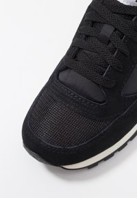 Baskets noires avec un mélange de suède et de tissu, affichant une surface texturée, un bout rond et une semelle intermédiaire blanche. Les lacets sont noirs et étroitement tissés.