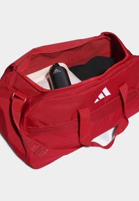 Bolsa deportiva roja con dos asas, exterior de tela, logo blanco en un lado, y contiene una botella de agua negra y ropa en su interior.