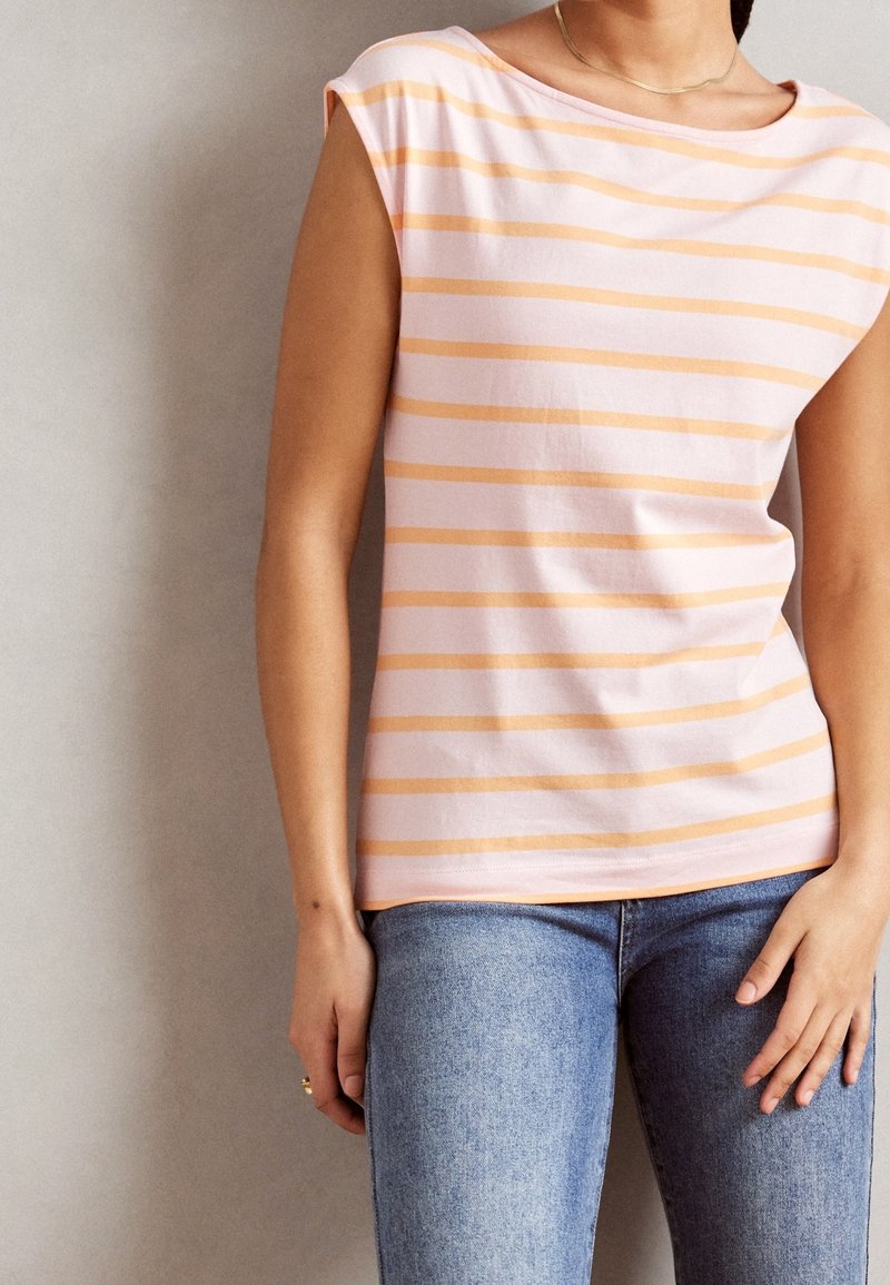 Femme portant un haut rose sans manches avec des rayures horizontales orange et un jean bleu, debout contre un mur clair uni.