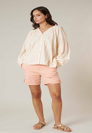 Femme portant un chemisier ample crème avec des motifs pêche, un short pêche, et des sandales beiges, debout devant un fond clair uni.