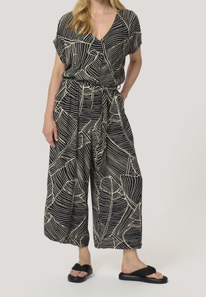 Femme portant un pantalon large noir et beige et un haut à manches courtes assorti avec un motif de feuilles audacieux, sandales noires, main dans la poche.