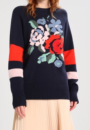 Pull bleu marine avec des rayures rouges et roses aux manches et un grand motif floral, porté avec une jupe plissée beige.