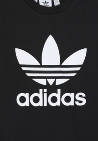 Černé bavlněné tričko s velkým bílým logem Adidas, se třemi paralelními pruhy nad názvem značky a designem listu v horní části.