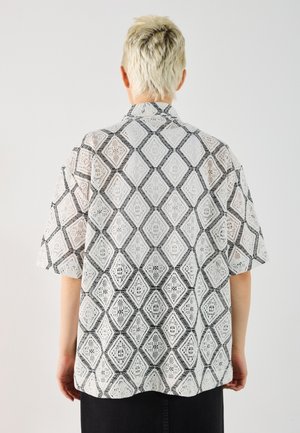 Persoon met kort blond haar, van achteren gezien, die een wit kanten hemd draagt met zwarte ruitpatronen en korte mouwen.