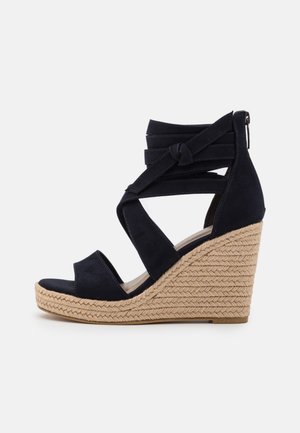 Marineblå ruskind kilehæl sandal med krydsede stropper, åben tå design og flettet jute kilehæl. Har en lynlåslukning bagpå.