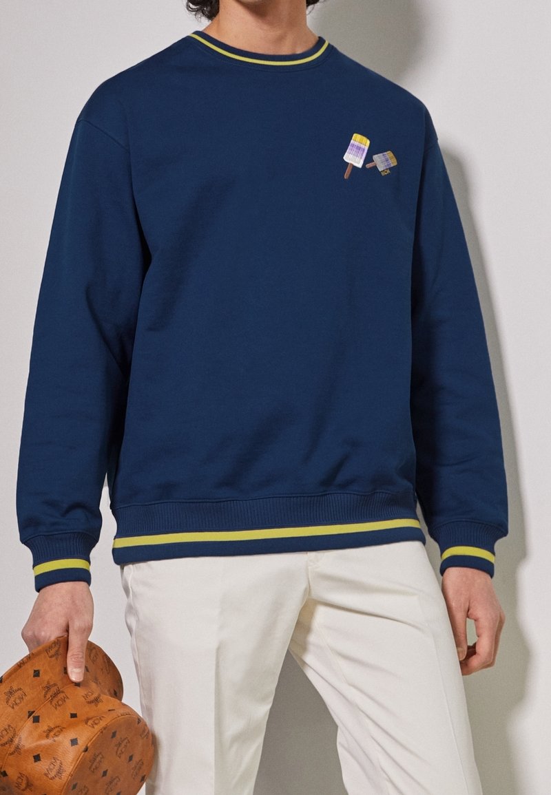 Sweat-shirt marine avec col et poignets côtelés accentués de jaune. Présente une broderie ludique de glace sur la poitrine. Texture de tissu lisse.