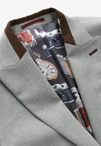 Blazer de espiga gris con cuello de ante marrón; el interior presenta un forro impreso con cámaras y relojes.