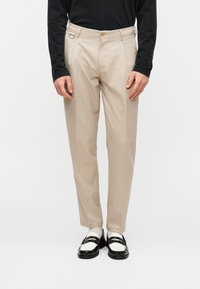 Pantalon beige ajusté avec un devant plat, une texture subtile, deux poches avant, un passant de ceinture et un pli central. Porté avec des mocassins noirs.