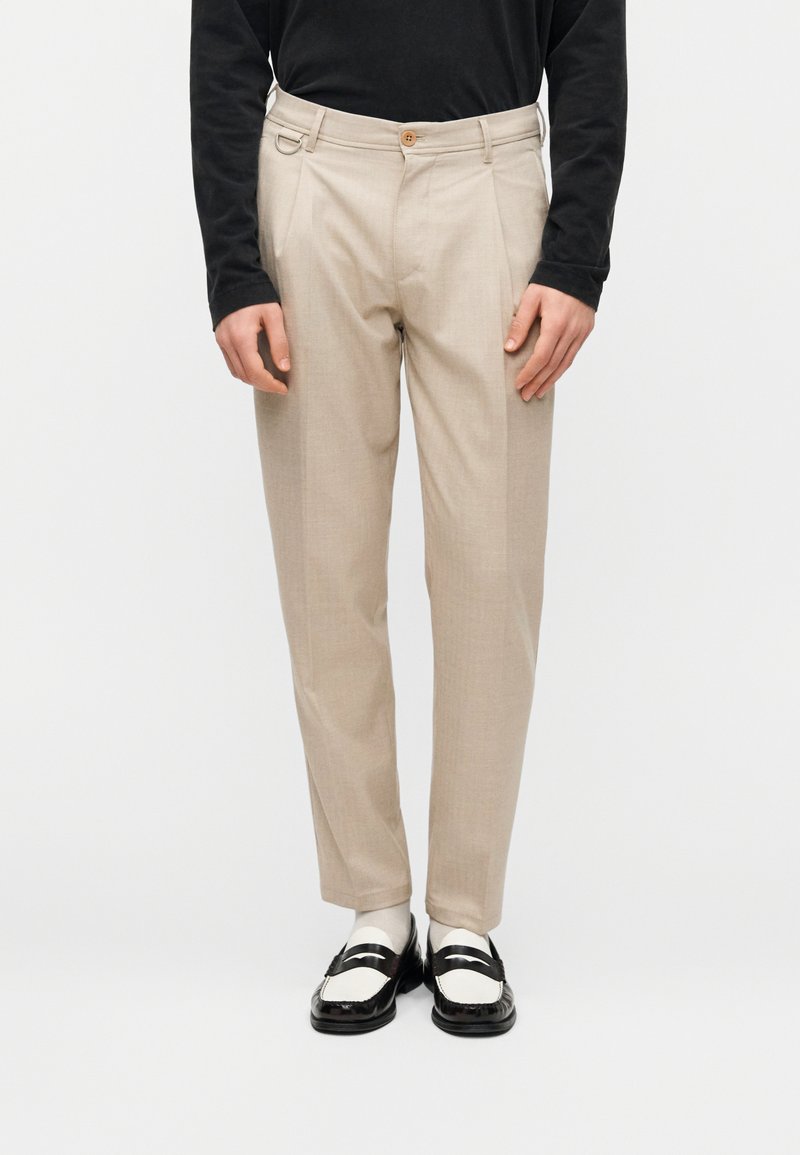 Pantalon beige ajusté avec un devant plat, une texture subtile, deux poches avant, un passant de ceinture et un pli central. Porté avec des mocassins noirs.