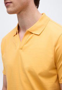 Homme portant un polo jaune à manches courtes avec un col ouvert, visible des épaules au menton sur un fond clair uni.