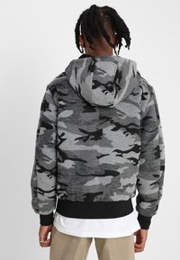 Sudadera con capucha de camuflaje en gris con detalles en negro, con un dobladillo y capucha ajustados. La tela parece suave con un acabado texturizado y aterciopelado.