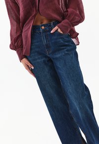 Burgunderfärgad genomskinlig blus med puffärmar, bärs över mörkblå high-waisted jeans med sidoklyftor och orangea sömnadsdetaljer.