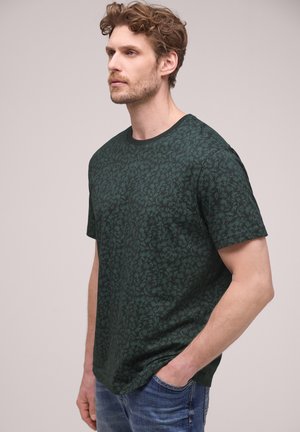 Mężczyzna o kręconych włosach, ubrany w ciemnozieloną, wzorzystą koszulkę i niebieskie dżinsy, stojący z jedną ręką w kieszeni na jednolitym tle.