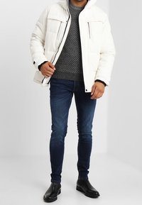 Veste blanche matelassée avec capuche, deux poches avant, pull gris texturé, jean bleu foncé et bottines noires. Design simple et décontracté.