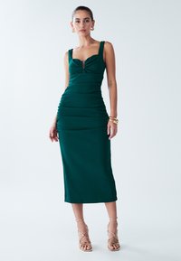 Teal fitted dress with a square neckline, gathered detailing at the waist, and thick straps. Smooth texture; knee-length. Paired with strappy heels.