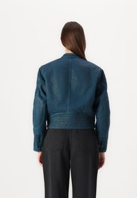 Chaqueta de mezclilla verde oscuro con un corte cropped y una cintura texturizada, que presenta acolchado en los hombros y puños acanalados, llevada sobre pantalones negros.