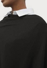 Sort strikket poncho med ribbet struktur, der har en lyseblå skjorte med krave nedenunder. Nærbillede fremhæver stoffets detaljer.