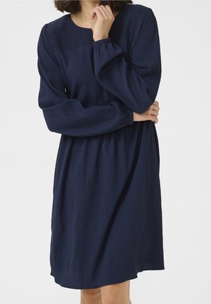 Femme portant une robe bleu marine texturée à manches longues, longueur genou, col carré et empiècement smocké.