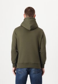 Olivengrønn hettegenser med stor, justerbar hette, ribbede mansjetter og midjan; brukt med blå jeans med en falmet tekstur og baklommer.