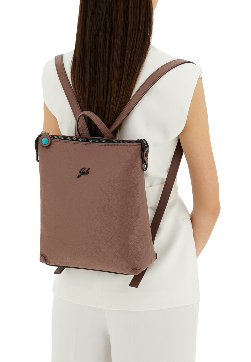 Zaino in pelle marrone con texture, spalline regolabili, chiusura con zip e un piccolo accento blu sul tirante della zip. Design minimalista.
