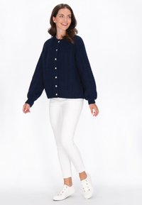 Cardigan a maglia a trecce blu navy con collo rotondo e bottoni argentati, abbinato a pantaloni bianchi slim-fit e sneakers bianche.