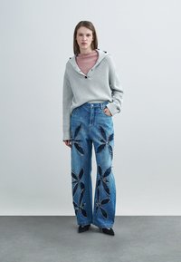 Pull gris côtelé avec capuche et détails à boutons, associé à un jean en denim bleu à jambes larges orné d'appliqués floraux en tissu foncé.