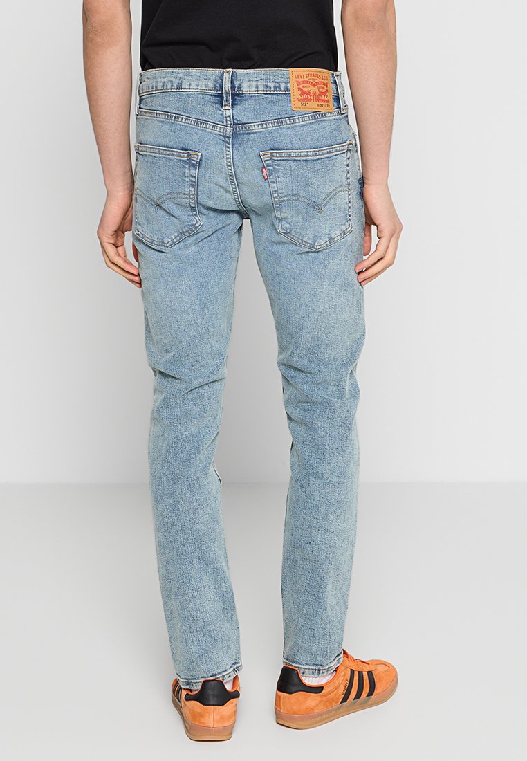 Persona vistiendo jeans ajustados Levi's 512 de color azul claro y zapatillas Adidas naranjas, de pie de espaldas contra un fondo blanco liso.