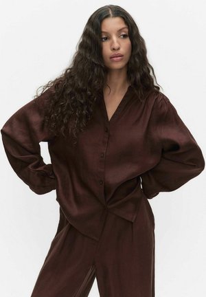 Kvinde med langt krøllet hår iført en løs mørkebrun skjorte med knapper og matchende bukser, stående med hænderne på hoften mod en hvid baggrund.