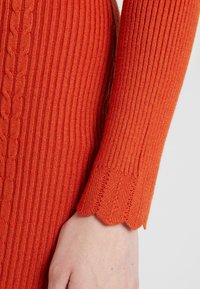 Texturerad orange tröja med ribbad design, kabelstickningsdetalj och scallopade manschetter. Nära bild av ärm och hand.