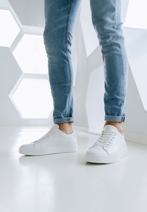 Sneakers bianchi a basse top in pelle liscia con punta arrotondata, design con lacci e una sottile suola a piattaforma. Indossati con jeans azzurri chiari con risvolto.