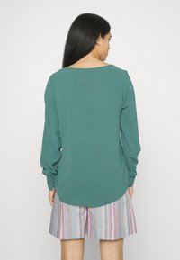 Top de manga longa em verde-água com decote redondo e punhos franzidos, combinado com calções às riscas multicolores, visto de costas.