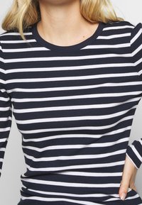 Haut à manches longues rayé bleu marine et blanc avec un col rond. Le tissu semble côtelé, présentant des rayures horizontales de largeur égale.