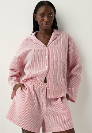 Kvinde iført rød og hvid langærmet skjorte med lodrette striber og matchende shorts med elastisk talje, stående med hænderne i lommerne.