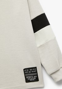 Langsleeve sporttop in lichtgrijze ribstof met zwart-witte strepen op de mouwen en een zwart label aan de zoom.