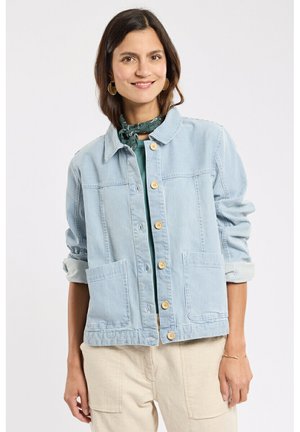 Femme souriante, portant une veste à rayures bleu clair avec des boutons en bois, un haut vert, un foulard vert autour du cou, un pantalon beige, et des boucles d'oreilles créoles dorées.