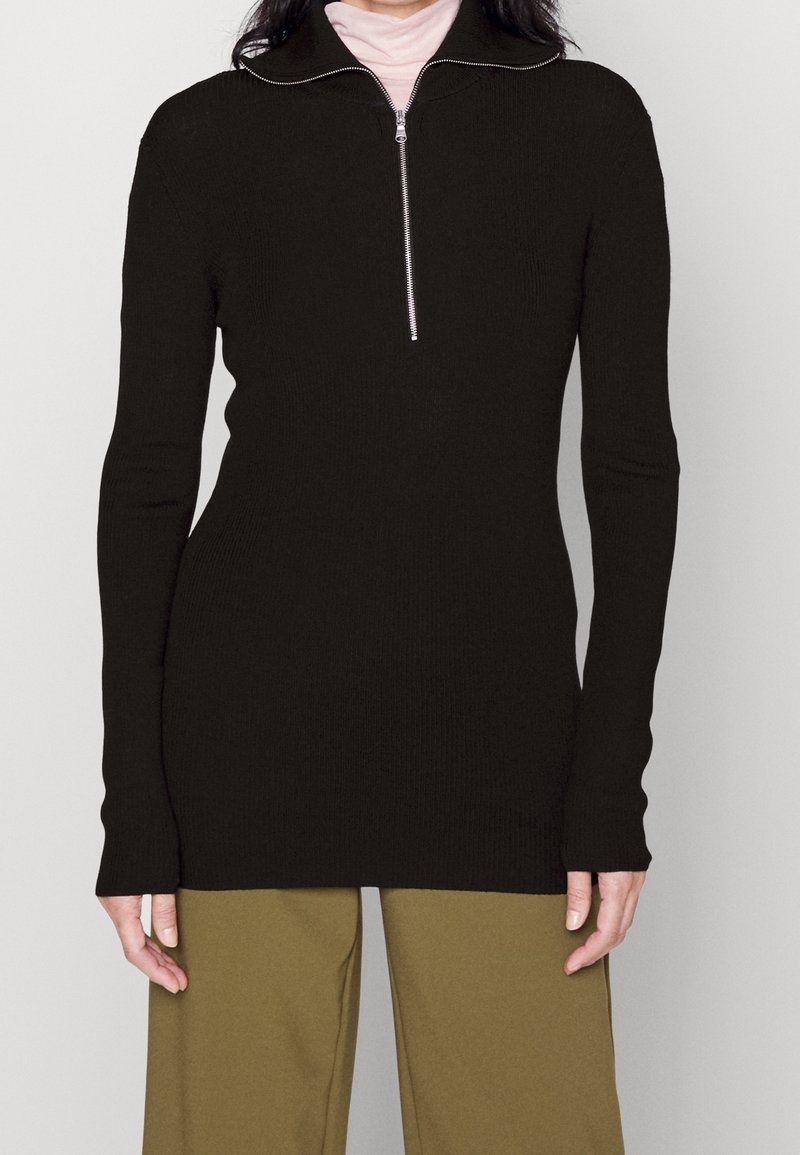 Svart ribbat långärmat topp med hög krage och dragkedja, med figursydda ärmar och en något förlängd kropp.