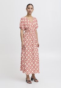 Abito maxi bianco con motivo geometrico rosso, maniche corte, scollo a V e vita con cordoncino. Indossato con sandali neri con tacco.