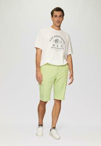 Jasnozielone szorty cargo z wieloma kieszeniami, zestawione z kremowym t-shirtem z grafiką, na którym widnieje napis "NYC" oraz widok na miejską panoramę. Białe sneakersy.