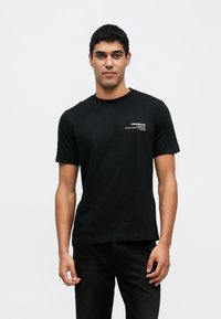 Czarna bawełniana koszulka z krótkimi rękawami, okrągłym dekoltem i subtelnym białym logo na lewej piersi. Noszona z ciemnymi dżinsami.