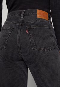 Vue arrière d'une personne portant un jean Levi's noir avec les poches arrière visibles, les passants de ceinture, et l'écusson en cuir de la marque Levi's sur la ceinture.