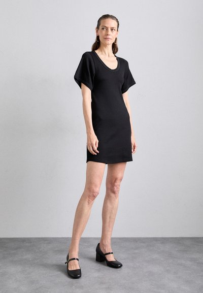 Schwarzes kurzärmeliges Kleid mit rundem Ausschnitt, aus weichem Material gefertigt. Hat eine gerade Passform und leichte Seitenschlitze. Kombiniert mit schwarzen Blockabsätzen.