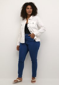 Chaqueta de denim blanca con cierre de botones y bolsillos con solapa, combinada con jeans skinny azul oscuro. Modelo lleva sandalias beige.