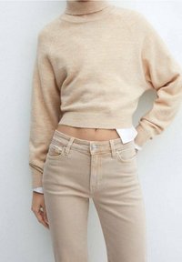 Pull court beige avec un col montant, texture douce ; associé à un jean taille haute beige clair, avec une fermeture simple par bouton et zip.